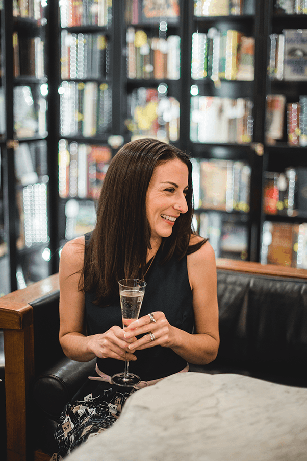About Me Portrait of Christine of Uncorked Asheville With Champagne Glass In A Bookstore