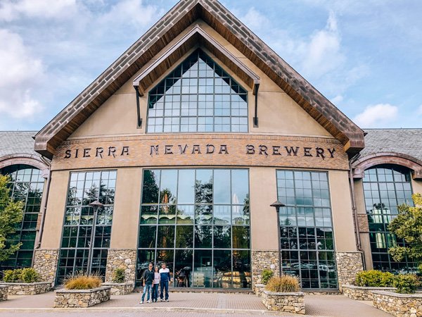 Tom and Christine's parents - two brunette white men and a white woman, standing outside of Sierra Nevada Brewing facility in Asheville, North Carolina; it is a large building with many windows